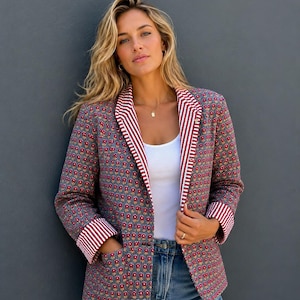 Pode incluir: Um blazer de manga comprida com base cinzenta e um padrão floral repetitivo em vermelho e amarelo. A peça possui lapelas e punhos forrados com tecido de riscas verticais brancas e vermelhas, dois bolsos frontais e um botão de fecho.