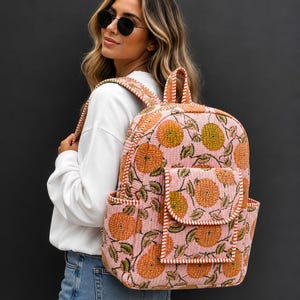 Puede incluir: Una mochila acolchada con base rosa claro y un patrón repetitivo de motivos florales circulares naranjas y hojas verdes. La bolsa incluye un bolsillo frontal con solapa, bolsillos laterales y correas con un borde a rayas naranjas y blancas.