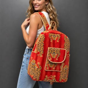May include: A bright red backpack featuring a repeating tiger pattern in black and gold. The backpack has a large main compartment, a smaller front pocket with a flap closure, and two adjustable shoulder straps with matching tiger print.