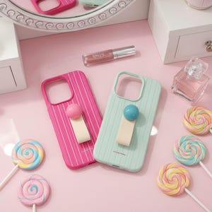May include: Two phone cases, one pink and one mint green, with vertical stripe patterns and a pop-up grip. The pink case has a pink ball grip, and the mint green case has a blue ball grip. The cases are on a pink surface with lollipops and beauty products.