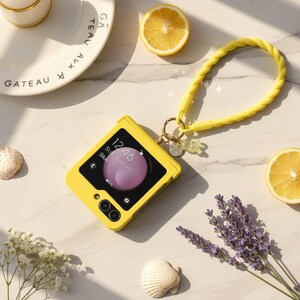 Op de afbeelding: Een gele smartphonehoes met een bijpassende polsband en een kleine bedel. Het telefoonscherm toont de tijd en datum. De afbeelding bevat ook citroenschijfjes, schelpen en lavendeltakjes.