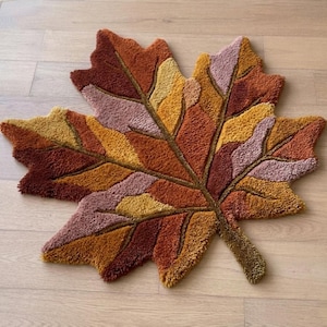 Puede incluir: Una alfombra con forma de hoja y diseño afelpado. Presenta colores otoñales: marrón, naranja, amarillo y rosa, dispuestos en un patrón de hoja. La alfombra está sobre un suelo de madera clara. Las dimensiones de la alfombra no son visibles.