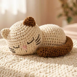 Op de afbeelding: Een gehaakte kattenknuffel in crème en bruin, liggend op een getextureerd crèmekleurig oppervlak. De kat heeft gesloten ogen, een roze neus en een bruin oor en staart. De handgemaakte kat is een zacht speelgoed.
