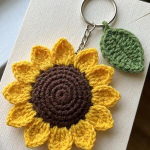 May include: A crochet sunflower keychain featuring a dark brown center and bright yellow petals. A small, textured green leaf is attached to the silver-toned metal keyring by a short chain, creating a floral accessory with distinct yarn stitching.