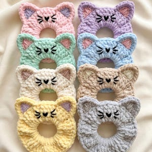 Op de afbeelding: Een verzameling van acht handgemaakte gehaakte scrunchies in de vorm van katten, in verschillende pastelkleuren. Elke scrunchie heeft geborduurde zwarte snorharen en een hartvormige neus. De kleuren zijn roze, paars, groen, blauw, crème, beige, geel en grijs.