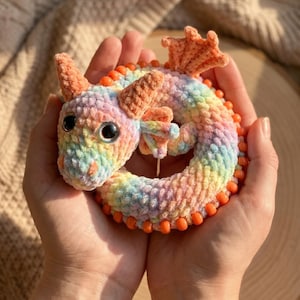 Puede incluir: Un juguete de dragón tejido a ganchillo en colores arcoíris, incluyendo rosa, amarillo, azul y naranja. El dragón tiene grandes ojos negros, cuernos naranjas y cuentas naranjas alrededor de su cuerpo. Un artículo hecho a mano caprichoso.