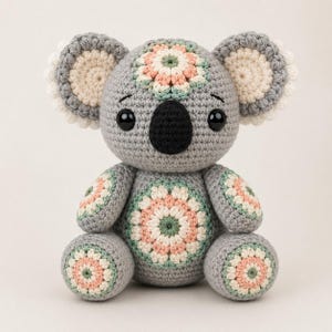 Può includere: Un koala all'uncinetto grigio fatto a mano con un naso nero e grandi occhi neri. Il koala presenta motivi floreali decorativi rosa, verdi e crema sulla testa, sul petto, sulle braccia e sui piedi. Le orecchie sono grigie con finiture color crema.