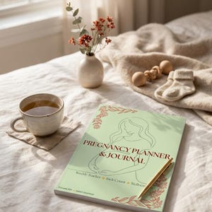Puede incluir: Un planificador y diario de embarazo verde claro con el título "Pregnancy Planner & Journal" y las palabras "Weekly Tracker + Kick Count + Wellness". Un bolígrafo dorado descansa sobre el diario. Una taza de té, un pequeño jarrón con flores y artículos para bebés también están en la imagen.