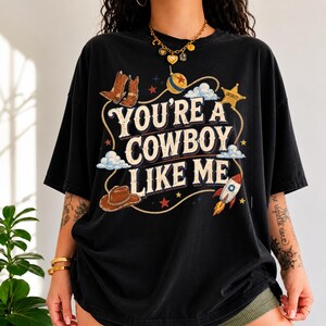 May include: A black short-sleeved t-shirt featuring a graphic print with the text "YOU'RE A COWBOY LIKE ME" in a vintage font. The design includes illustrations of cowboy boots, a hat, a sheriff star, a rocket, and a lasso rope.