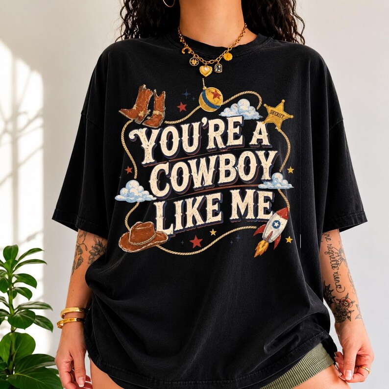 May include: A black short-sleeved t-shirt featuring a graphic print with the text "YOU'RE A COWBOY LIKE ME" in a vintage font. The design includes illustrations of cowboy boots, a hat, a sheriff star, a rocket, and a lasso rope.