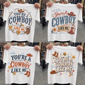 May include: A white t-shirt featuring a graphic print with the text "YOU'RE A COWBOY LIKE ME" in various fonts. The design includes illustrations of a cowboy hat, a rocket ship, a sheriff star, a lasso, a ball, and cowboy boots in colors.