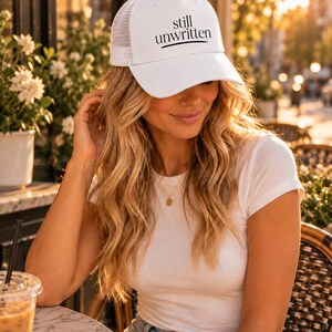 May include: A white trucker cap featuring a mesh back and a solid front panel. The text "still unwritten" is printed in black on the front of the cap, with a horizontal black line underneath the text. The cap has a curved brim and a structured crown.
