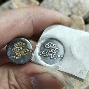 Pode incluir: Close-up de dois objetos de metal prateado com um desenho de nuvem. Um é um pequeno pino redondo com um desenho de nuvem dourada. O outro é uma peça retangular maior com um desenho de nuvem semelhante. O fundo está desfocado.
