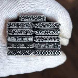 May include: A collection of ten rectangular metal stamping tools arranged in a stack. Each tool features a unique engraved pattern on its face, including various scrollwork, geometric Greek key designs, chevron lines, and leaf-like motifs.