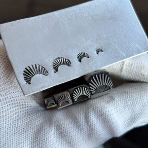 Peut inclure: Un ensemble de quatre outils de frappe en métal à tige carrée, chacun présentant un motif en éventail ou en rayon de soleil à l'extrémité. Les outils sont présentés avec une plaque métallique rectangulaire portant quatre empreintes du motif.