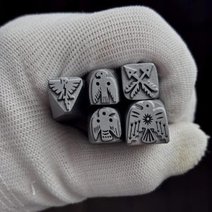 Puede incluir: Un conjunto de cinco herramientas de estampado de metal en tono plateado, cada una con un diseño grabado distinto en su cara. Los diseños incluyen un ave estilizada con las alas extendidas, un ave con un símbolo solar y dos flechas cruzadas, todo con un acabado mate.