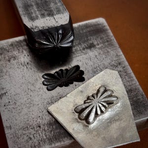 Puede incluir: Una herramienta de estampado de metal con diseño de estrella, un bloque de metal cuadrado con una impresión de estrella estampada y una pieza triangular de metal plateado con un patrón de estrella en relieve.
