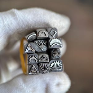 May include: A collection of twelve square metal stamps arranged in a grid, each featuring a unique engraved design including sunbursts, geometric triangles, fan shapes, and radiating lines, all crafted from a dark, matte-finished metal material.