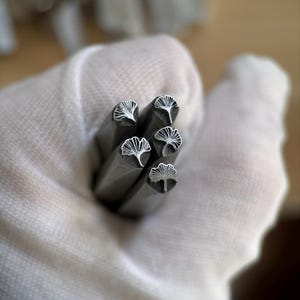 Può includere: Primo piano di cinque timbri metallici con disegni a forma di foglie di ginkgo. I timbri sono tenuti in una mano guantata di bianco. I disegni delle foglie di ginkgo sono incisi nel metallo. I timbri sono grigi e rettangolari.