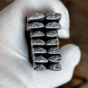 May include: A collection of twelve silver-toned metal stamping tools arranged in two columns. Each tool features a unique engraved design on its face, consisting of various mountain range silhouettes on the left and stylized ocean wave patterns on the right.