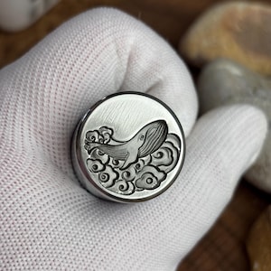 May include: A circular silver-toned metal wax seal stamp featuring an engraved design of a whale swimming amidst stylised clouds. The stamp has a brushed metallic finish and a smooth, flat circular face with deep relief detailing of the whale.