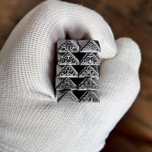May include: A set of ten silver-toned metal stamps arranged in two columns, each featuring a unique, intricate triangular filigree pattern. These metal tools are designed for embossing or marking surfaces with detailed, decorative corner designs.