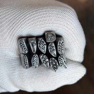 May include: A collection of eleven silver-toned metal stamping tools, each featuring a distinct botanical design engraved on the flat end. The designs include various leaf patterns, sprigs, and a small cluster of three flowers, all arranged together.