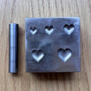 May include: A square metal dapping block featuring five heart-shaped indentations of varying sizes, accompanied by a single cylindrical metal punch tool. The surface of the block shows a brushed metallic finish with visible tool marks.