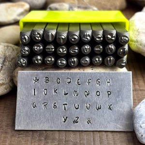 Peut inclure: Un ensemble de tampons de lettres en métal avec un étui de rangement jaune vif. Les tampons sont disposés au-dessus d'une plaque métallique avec l'alphabet estampé. Les lettres sont de style manuscrit. Le fond est une surface en bois avec des pierres.