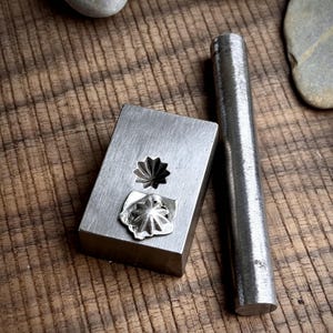 May include: A metal jewelry making tool set. The set includes a rectangular die with two floral impressions and a cylindrical metal punch. The die and punch are made of silver-colored metal and sit on a wooden surface.