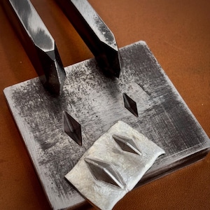 May include: A square metal block with a brushed finish featuring two diamond-shaped indentations, positioned alongside two matching metal punches with pointed tips and a small rectangular metal plate showing two raised diamond-shaped impressions.