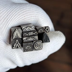 Puede incluir: Un conjunto de diez pequeñas herramientas de estampado de metal con extremos cuadrados que presentan varios diseños grabados, incluyendo patrones geométricos, un sol, un engranaje y un arco curvo, agrupados con un acabado gris metálico texturizado.