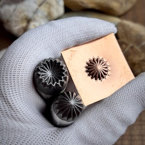 Könnte beinhalten: Nahaufnahme von Metallhandwerkzeugen. Zwei Metallstempel mit einem floralen Design werden neben einem Kupferquadrat mit dem gleichen Design gehalten. Die Werkzeuge werden in einem weißen Handschuh gehalten.