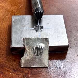 Op de afbeelding: Een metalen stempelgereedschap met een waaiervormige punt boven een rechthoekig stalen blok. Het blok heeft een centraal ingedrukt waaierontwerp. Een dun, onregelmatig zilverkleurig metalen stukje ervoor toont een duidelijke, verhoogde waaierafdruk.