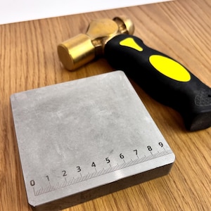 Könnte beinhalten: Ein quadratischer Metall-Ambossblock mit einem aufgedruckten Lineal an der Unterkante, das bis zu 9 Zentimeter misst, neben einem Hammer mit Messingkopf, schwarzem Griff mit gelben Akzenten und einer strukturierten Grifffläche am Ende.