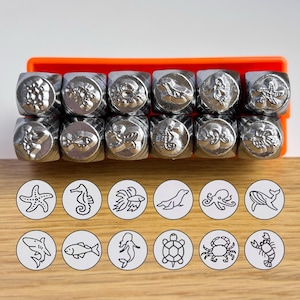 May include: A set of twelve silver-toned metal stamps arranged in two rows, each featuring a unique embossed marine-themed design. The collection includes icons of a starfish, seahorse, fish, turtle, octopus, crab, lobster, whale, shark, seal, and mermaid, displayed with corresponding black ink impressions.