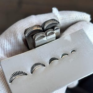 Op de afbeelding: Een set van vier metalen stempelgereedschappen met gebogen, zonnestraalachtige patronen aan de uiteinden, samen met een rechthoekige metalen plaat met vier verschillende afdrukken van hetzelfde zonnemotief in verschillende groottes.