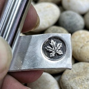 May include: A silver metal money clip with a rectangular shape and a circular emblem featuring a detailed maple leaf design. The clip is held against a background of smooth, rounded stones.