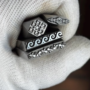 Puede incluir: Un conjunto de cuatro herramientas de estampado de metal con distintos patrones grabados. Una herramienta presenta una cuadrícula geométrica, otra una filigrana alargada, una un diseño de ondas continuas y un motivo de enredadera floral.