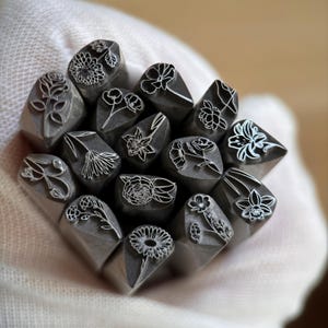 May include: A collection of metal stamps with various floral designs. Each stamp features a different flower, including roses, daisies, and other detailed botanical illustrations. The stamps are held in a white gloved hand.