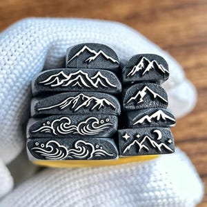 May include: A collection of eight metal stamping tools featuring engraved designs. The stamps display various mountain ranges, stylized ocean waves, and a night scene with mountains, stars, and a crescent moon, all rendered in white on dark metal.