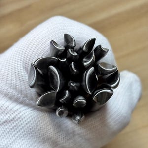 May include: A cluster of metallic, dark grey, pointed cylindrical tools arranged in a circular formation. Each individual tool features a curved, concave tip with a polished finish, creating a dense, textured, and multi-faceted appearance.