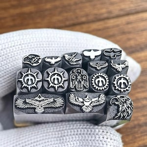 Peut inclure: Un ensemble de quinze outils de frappe en métal présentant divers motifs d'aigles et d'oiseaux. Chaque outil possède une tête carrée ou rectangulaire avec un motif gravé distinct, incluant des têtes d'aigle détaillées, des ailes et des soleils.