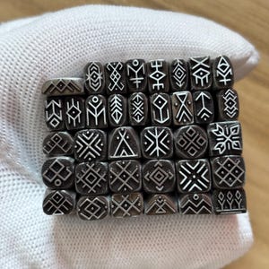 May include: A collection of small, rectangular metal stamps arranged in a grid. Each stamp features a unique, engraved geometric symbol or line pattern in a dark, oxidized silver finish. The stamps are uniform in size and shape, displaying various intricate designs including diamonds, crosses, and abstract linear motifs.