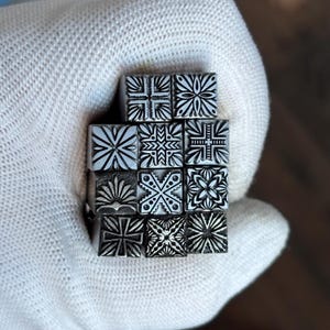 Puede incluir: Un conjunto de once sellos metálicos cuadrados pequeños dispuestos en una cuadrícula, cada uno con un patrón geométrico o floral grabado único. Los sellos son de color plata metálica con líneas oscuras y profundas que resaltan los diseños.