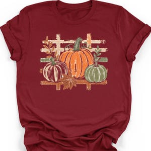 May include: A maroon short-sleeved t-shirt featuring a central graphic of three pumpkins in orange, muted red, and sage green. The design includes a distressed plaid background pattern, autumn leaves, and the word MAROON printed at the bottom.