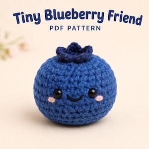 May include: A blue crocheted blueberry with a dark blue top, black safety eyes, a stitched smile, and pink cheeks. The text above reads Tiny Blueberry Friend PDF PATTERN. The blueberry is round with a textured yarn surface and a small stem area.