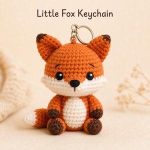 May include: A crocheted fox keychain in orange, white, and brown. The fox has a round body, large black eyes, and a small black nose. The keychain has a silver ring attached to the top of the fox's head. The text "Little Fox Keychain" is above the fox.
