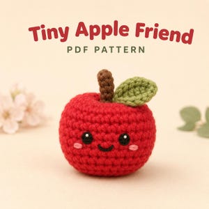 May include: A red crocheted apple toy with a brown stem and a green leaf. The apple features embroidered black eyes, a small black smile, and pink cheeks. Above the apple, the text reads Tiny Apple Friend PDF PATTERN in red and green.