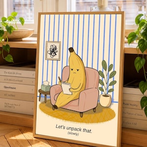 May include: A framed wall art print featuring a yellow banana character sitting in a pink armchair, holding a notepad and pen. The background has blue vertical stripes, a small framed drawing, a tissue box, and a potted plant. Text reads: Let's unpack that. (slowly).